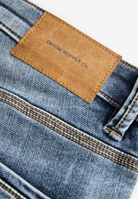Džínsy z denimu s hnedou koženou nášivkou s nápisom "DENIM SUPPLY Co." Tkanina je modrý denim s oranžovým prešívaním a textúrovaným povrchom.