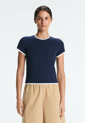 Femme aux cheveux foncés portant une chemise bleu marine à manches courtes avec un liseré blanc, se tenant devant un fond clair uni, portant un pantalon beige.