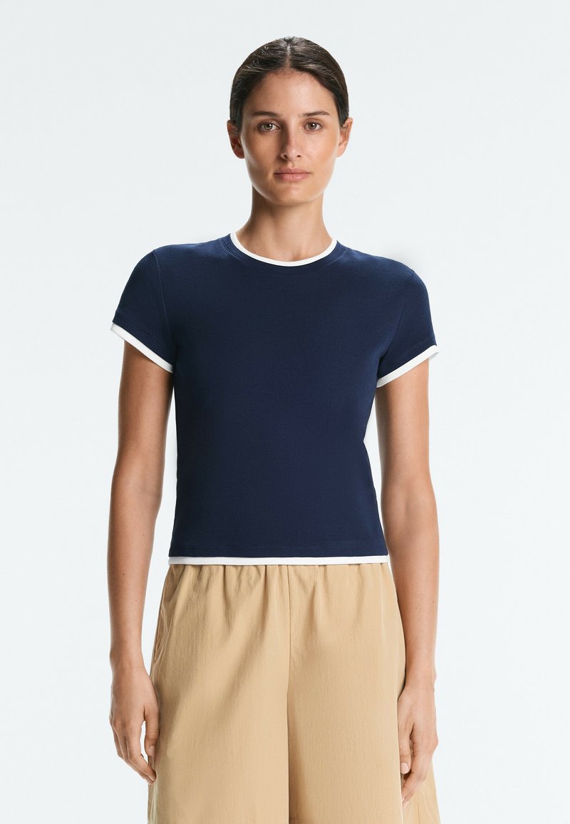 Femme aux cheveux foncés portant une chemise bleu marine à manches courtes avec un liseré blanc, se tenant devant un fond clair uni, portant un pantalon beige.