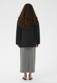 Črna prešita jakna s pravim krojem, kombinirana z dolgo sivo krilo z vzorcem in ravnimi črnimi sandali, pogled od zadaj.