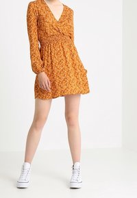 Pitkähihaine kietaisumekko oranssina, jossa on pieniä kukkakuoseja, varustettuna smokkivyöllä, polven ylle ulottuvalla pituudella, ja yhdistetty valkoisiin lenkkareihin.