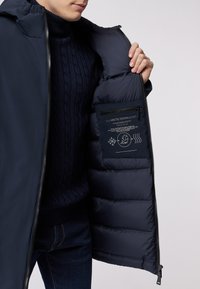 Marineblaue Daunenjacke mit geripptem Kragen, gestepptem Innenfutter und Reißverschluss-Innentasche, die Pflegehinweise und Produktinformationen zeigt.