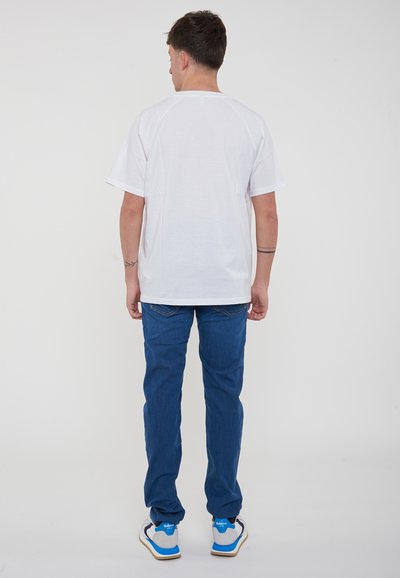 T-shirt bianca in cotone con maniche corte e vestibilità giovane, abbinata a jeans in denim blu e sneakers grigie con accenti blu.