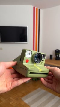 Green instant camera with a rectangular shape, featuring a lens, two flash bulbs, and a red button. Notable yellow stripe and textured surface.