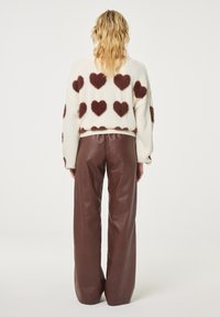 Oversized cream sweater with fuzzy brown heart patterns, paired with high-waisted brown leather trousers, featuring a smooth texture and wide leg.