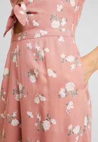 Nahaufnahme eines rosa Blumenkleides mit weißen Blüten, das eine gebundene Schleifendetail und eine Hand zeigt, die in einer seitlichen Tasche ruht.