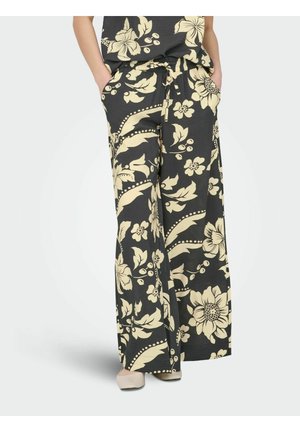 Czarne spodnie o luźnym kroju z dużym beżowym kwiatowym wzorem, ściągaczem w pasie i bocznymi kieszeniami, noszone z beżowymi butami, ręce w kieszeniach.