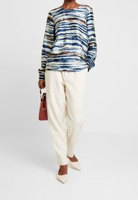 Färgglad långärmad topp med blå, vit och brun horisontella ränder, i kombination med krämfärgade tapered byxor och beige spetsiga skor.