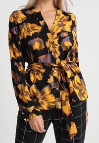Långärmad svart blus med stort mönster av gula och lila blommor, v-ringning och knytbälte vid midjan, med knappdetaljer.