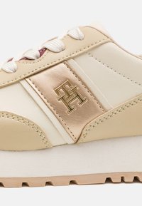 Sneaker in pelle beige e crema con una striscia e logo dorati, superficie texture e lacci bianchi. Suola spessa e leggera.