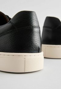 Adidași negri din piele cu o finisare texturată, având o talpă din cauciuc crem și un vârf cu model în romb pentru o aderență suplimentară.
