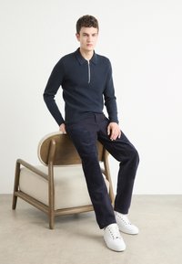Marineblauwe trui met rits rond de hals, ribbelstructuur, gecombineerd met een donkere corduroy broek met patchaccenten. Witte sneakers maken de outfit compleet.