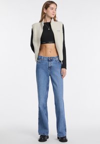 Fleece-Weste in Creme über einem schwarzen Cropped-Top, kombiniert mit blauen hochgeschnittenen Jeans und schwarzen Plateaustiefeln. Einfaches Design mit lässiger Passform.