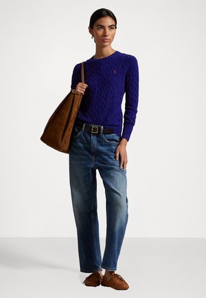 Lila Kabelstrickpullover, blaue Jeans, braune Wildledertasche und Mokassinschuhe. Der Pullover hat gerippte Bündchen und ein kleines Logo.
