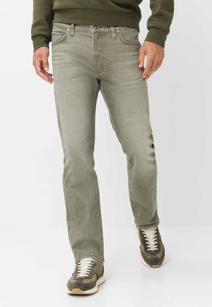 Mann trägt olivgrüne Slim-Fit-Jeans, dunkelgrünen Pullover und grün-weiße Sneakers und steht vor einem schlichten Hintergrund.