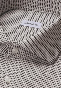 Camicia con colletto, motivo a diamante nero e beige e bottoni bianchi, dotata di un'etichetta in stoffa bianca con il testo "seidensticker" all'interno del colletto.