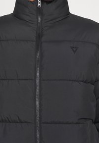 Giacca in piuma nera con colletto alto, chiusura con zip, design trapuntato e logo triangolare in rilievo sul petto sinistro. Texture morbida.
