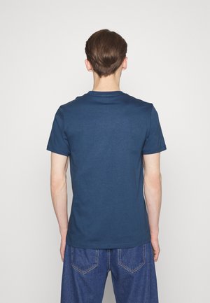 Mann mit kurzen braunen Haaren, der ein schlichtes dunkelblaues T-Shirt mit kurzen Ärmeln und blaue Jeans trägt, von hinten vor einem weißen Hintergrund.