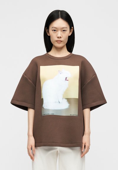 Γυναίκα με ίσια μαύρα μαλλιά φοράει φαρδιά καφέ πουκάμισο με εκτύπωση λευκής χασμουρητής γάτας σε μπεζ φόντο, συνδυασμένο με λευκό παντελόνι.