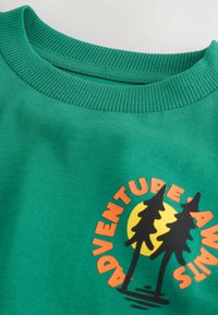 Zöld pamut pulóver bordás gallérral. Két fát ábrázoló grafikai tervezés található rajta, valamint a "KALAND VÁR RÁ" felirat narancssárga és fekete színben.