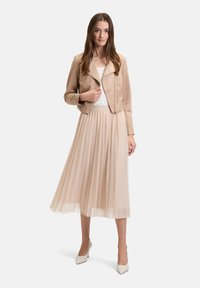 Giacca cropped in suede beige con cerniera, abbinata a una gonna midi plissettata di colore crema e scarpe con tacco alto bianche. Texture liscia, stile aderente.