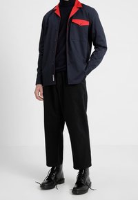 Mörkblå skjorta med knäppning och röd krage samt bröstficka, tillsammans med svarta vida byxor och svarta snörstövlar.