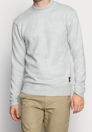 Mann trägt hellgrauen Rippstrickpullover und beige Hose, steht mit entspannten Armen seitlich an einem einfarbigen Hintergrund.