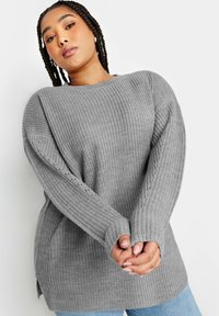Grå stickad tröja med ribbad textur, långa ärmar med sidoslitsar och en avslappnad passform, ihop med ljusblå jeans.
