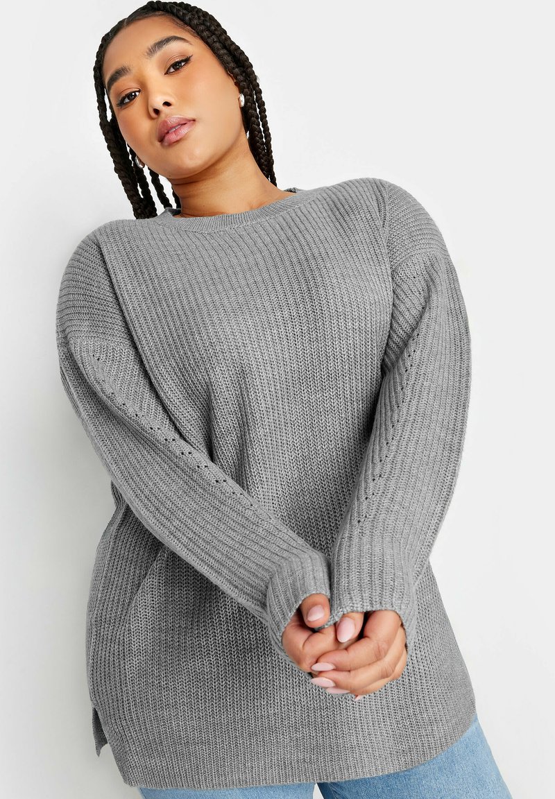 Grå stickad tröja med ribbad textur, långa ärmar med sidoslitsar och en avslappnad passform, ihop med ljusblå jeans.