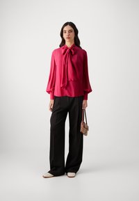 Fuchsia bluse med en stor sløjfe, elastiske manchetter, parret med sorte vide bukser og cremefarvede slip-on sko, mens hun holder en lille håndtaske.