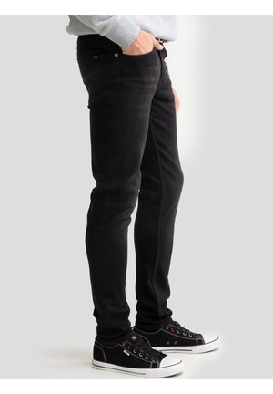 Osoba nosząca czarne jeansy o dopasowanym kroju i czarne niskie trampki z białą podeszwą, stojąca z jedną ręką w kieszeni na jednolitym tle.