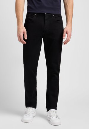 Mand iført sorte slim fit jeans, hvide sneakers og en mørk skjorte, stående mod en lys grå ensfarvet baggrund.