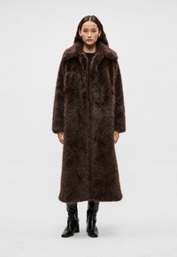 Hnedý dlhý kabát z umelého kožúška so širokým golierom, nadrozmerným strihom a textúrovanou látkou. Modelka má na sebe priliehavé čierne čižmy.