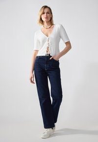 Haut blanc texturé avec un décolleté en V profond et un laçage à l'avant, associé à un jean taille haute bleu foncé et des baskets blanches.