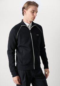 Sort zip-up jakke med hvide pipings detaljer, lavet af blødt stof. Har sidelommer og et logo på brystet.