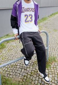 Chaqueta negra y morada con acentos blancos, camiseta blanca de los Lakers con gráficos en morado y amarillo, pantalones negros con ribete blanco y zapatillas.