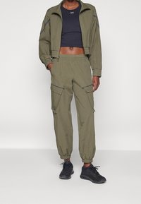 Person wearing olive green cargo pants and matching cropped jacket, black crop top, and black athletic sneakers standing against white background.