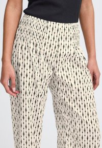 Pantalon léger de couleur crème avec un motif flèche noire. Il possède une taille élastique et des poches latérales. Texture en coton lisse.