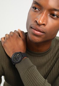 Montre avec bracelet en silicone noir et cadran noir texturé ; présente des index horaires dorés et un affichage de la date. Portée au poignet d'une personne.