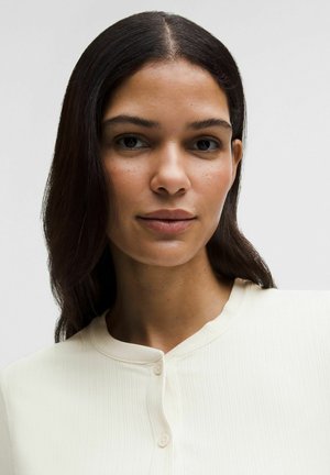 Młoda kobieta o długich ciemnych włosach, ubrana w kremową bluzkę na guziki, patrząca prosto w obiektyw na tle jednolitego, jasnego tła.