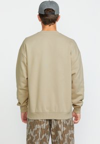 Sudadera beige con cuello redondo y hombros caídos, combinada con pantalones de camuflaje en tonos marrón y beige con patrones. Vista trasera, sin logotipo visible.