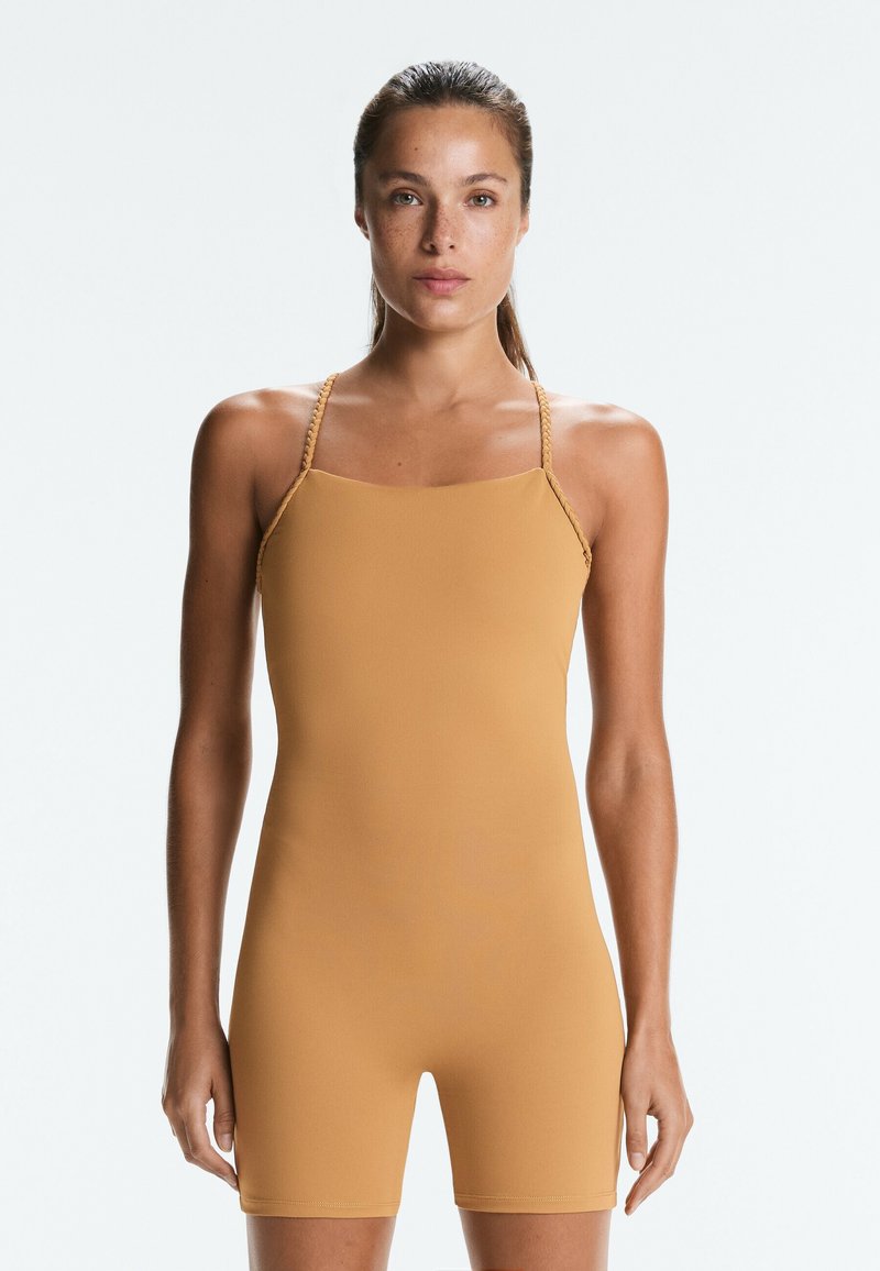 Femme aux cheveux attachés porte une combinaison une-pièce sans manches, beige, avec des bretelles tressées, debout devant un fond clair uni.