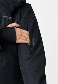 Hand placing on black jacket arm with zippered pocket labeled "STX 20.000" and partial "Swedemount" text on chest area.