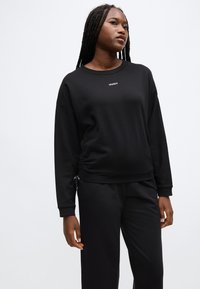 Črni pulover z dolgimi rokavi, z okrolim izrezom in nabranim stranskim detajlom. Logotip "HUGO" v majhnih belih črkah na prsih.