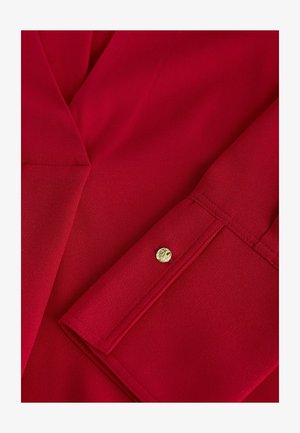 Rote Bluse mit glatter Textur, ausgestattet mit einem Reverskragen und einem detailreichen Knopfaufschlag mit goldenen Akzenten.