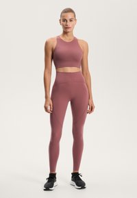Atletski komplet v prašno roza barvi, ki vključuje kratko majico in legice z visokim pasom. Gladka, raztegljiva tkanina s konturnimi šivi, v kombinaciji s črnimi supergami.
