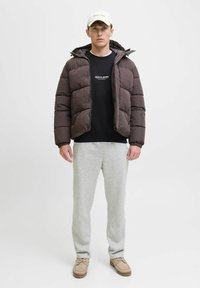 Bruine pufferjack met een capuchon, zwarte sweatshirt, grijze trainingsbroek en beige schoenen. De jas heeft een gewatteerd ontwerp met een rits.