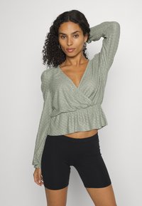 Top de manga longa em verde-claro, com padrão texturizado, decote em V e bainha com babados. Combinado com calções pretos de cintura alta.