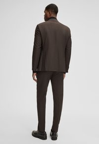 Brauner Anzug mit strukturiertem Design, ausgestattet mit einem einzelnen Schlitz und schmal geschnittenen Hosen, kombiniert mit schwarzen Schuhen. Glatte Stoffstruktur.