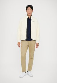 Flis jakna u off-white boji s crnim patentnim zatvaračem i detaljima, uparena s bež cargo hlačama i bijelim tenisicama. Model stoji pred jednostavnom pozadinom.